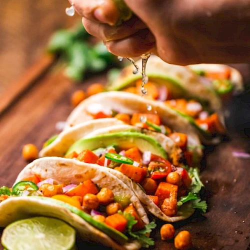 A hand squeezes lime over a row of vibrant tacos filled with chickpeas, avocado, tomatoes, and greens on a wooden board.