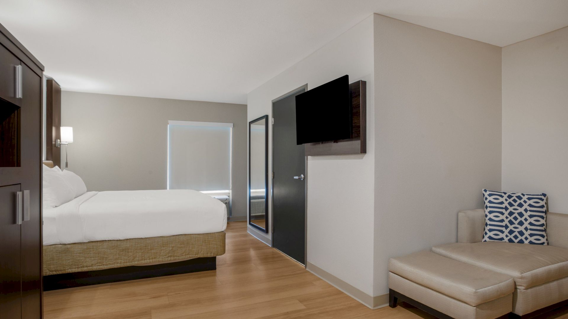 The image shows a modern hotel room with a bed, wall-mounted TV, sofa, and wooden floor, featuring a neutral color palette and simple decor.