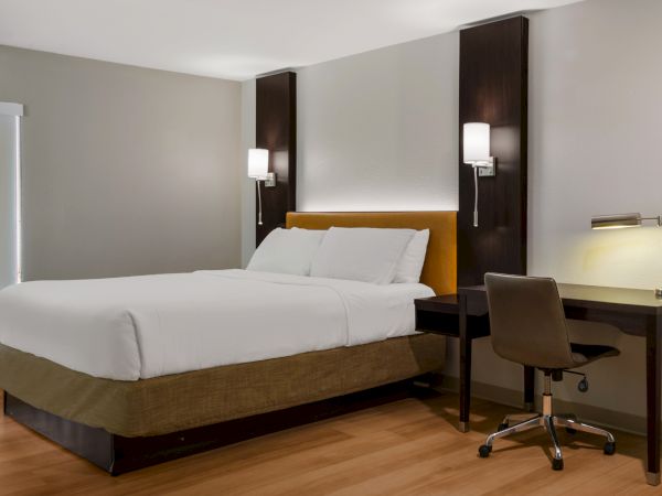 A modern hotel room with a neatly made bed, two bedside lamps, and a desk with a chair and lamp.