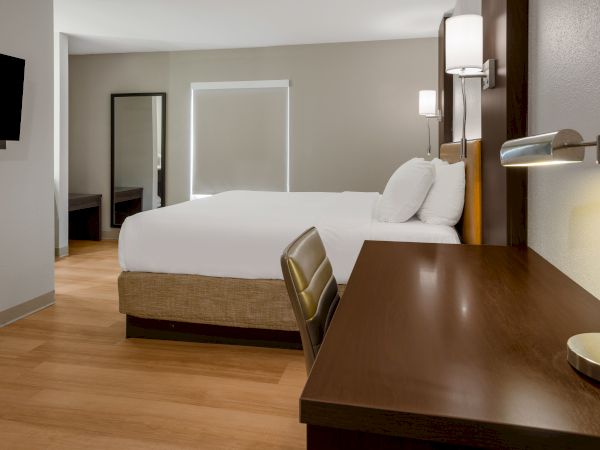 A modern hotel room with a double bed, desk, chair, TV, and mirror, featuring minimalist decor and wooden flooring.