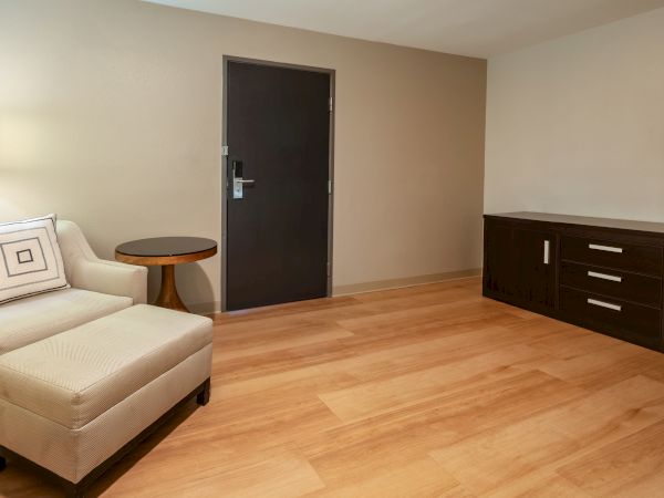 The image shows a room with a beige armchair, a round side table, a dark door, wooden flooring, and a dark cabinet with a light countertop.