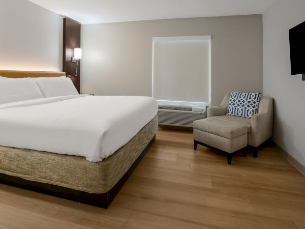 The image shows a modern hotel room with a large bed, a chair, a small table, and a wall-mounted TV.