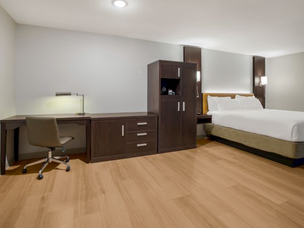 The image shows a modern hotel room with a bed, desk, chair, and wardrobe. Wooden flooring and neutral walls create a minimalist look.