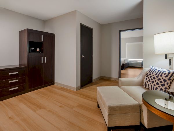 A modern room with wooden flooring, a sofa, a lamp on a side table, a cabinet, and an open door showing a glimpse of a bedroom.