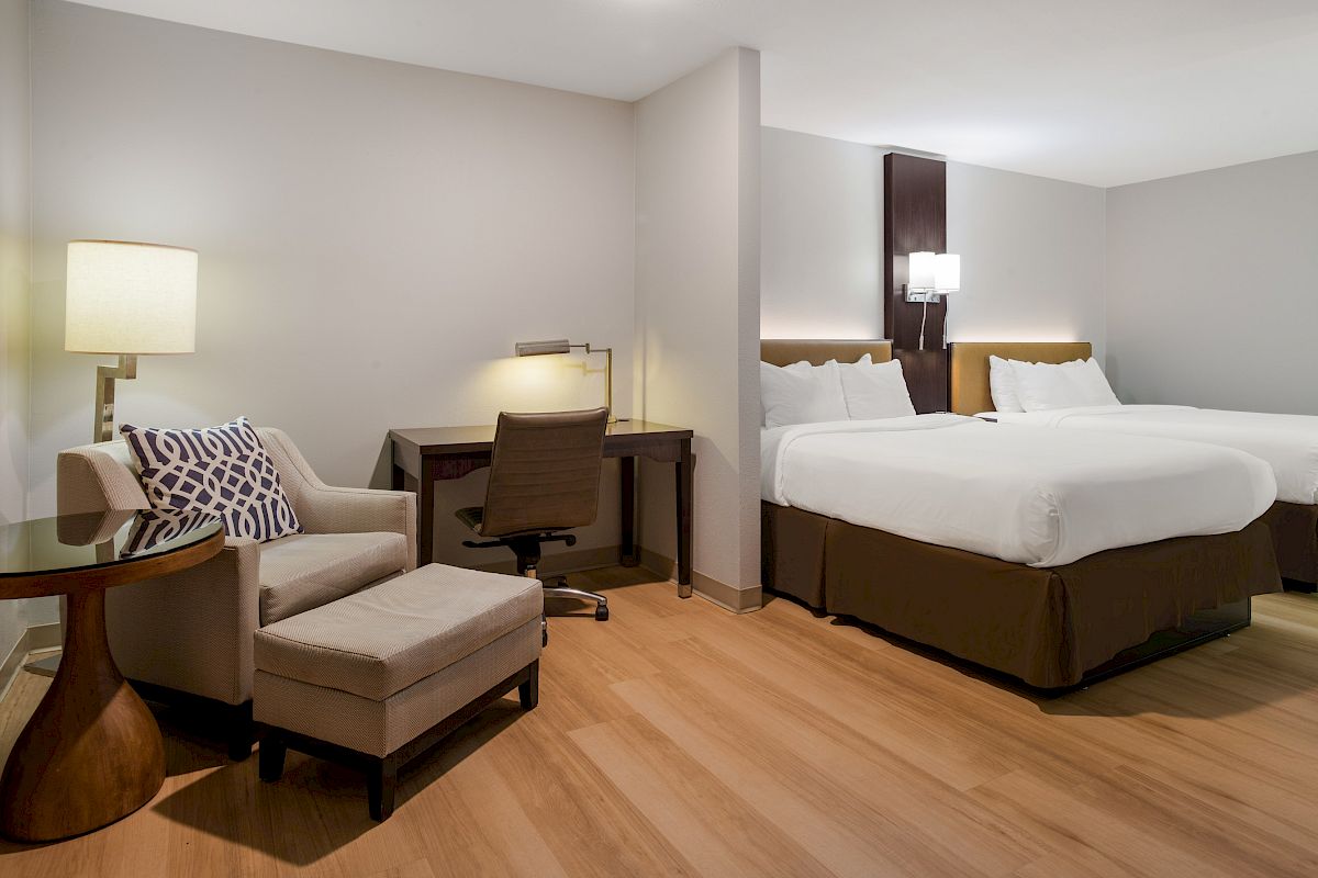 The image shows a modern hotel room with two beds, a desk with a chair, and a cozy seating area featuring an armchair and ottoman.