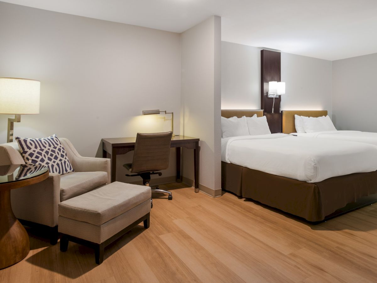 The image shows a modern hotel room with two beds, a desk with a chair, and a cozy seating area featuring an armchair and ottoman.