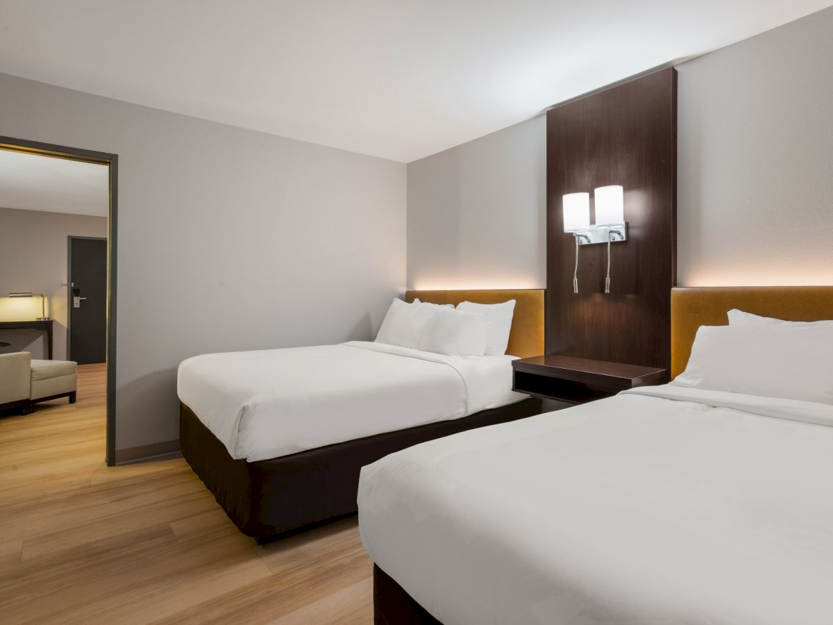 A hotel room with two double beds, a wooden accent wall, modern lighting, and an adjoining living area with a sofa and lamp.