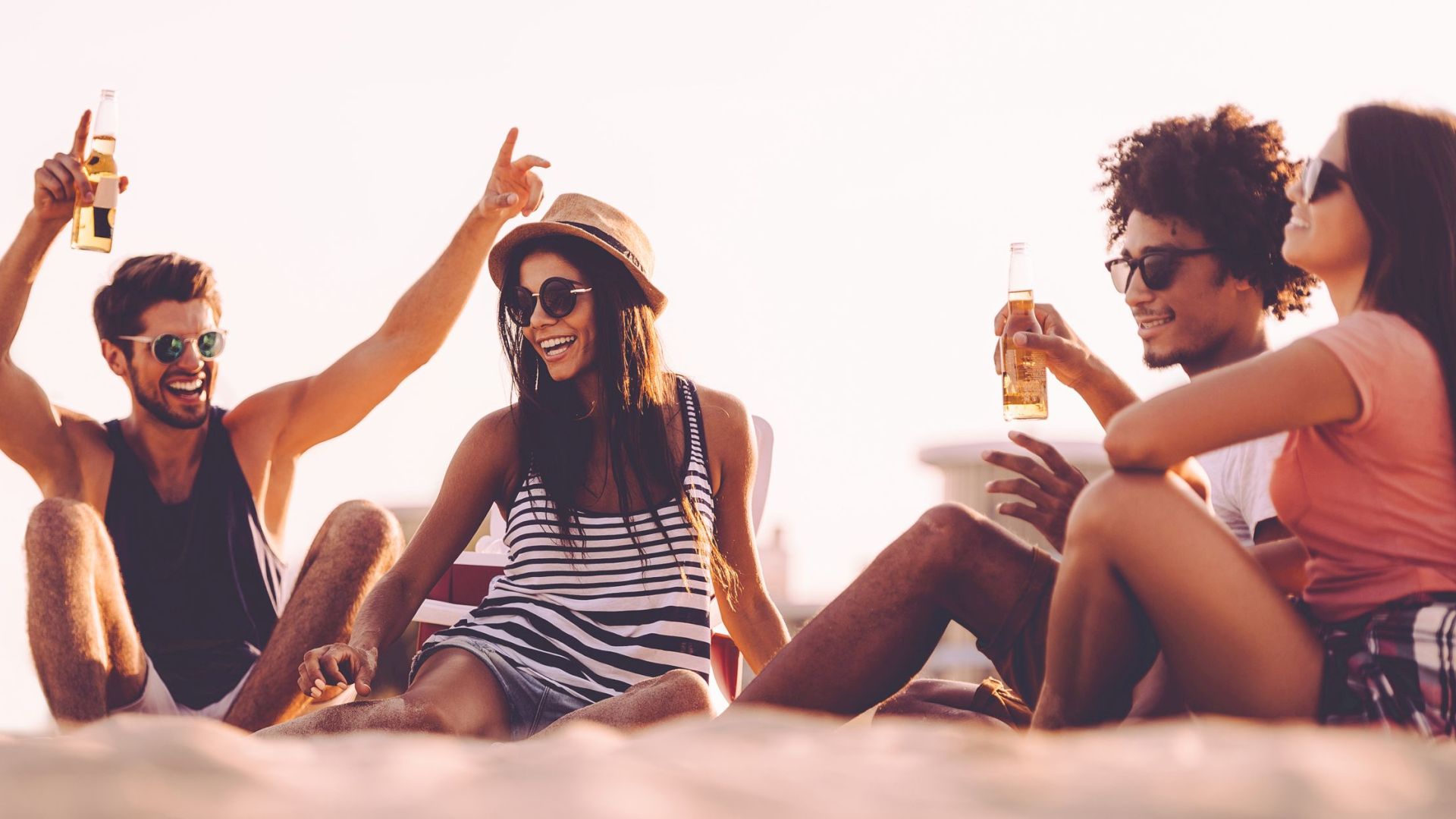 A group of four friends is sitting on a beach, enjoying drinks and laughing together in a relaxed, sunny setting.